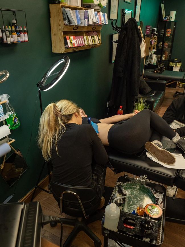 A tattoo artist works on a person lying on a studio table, illuminated by a ring light in a dark-walled shop.