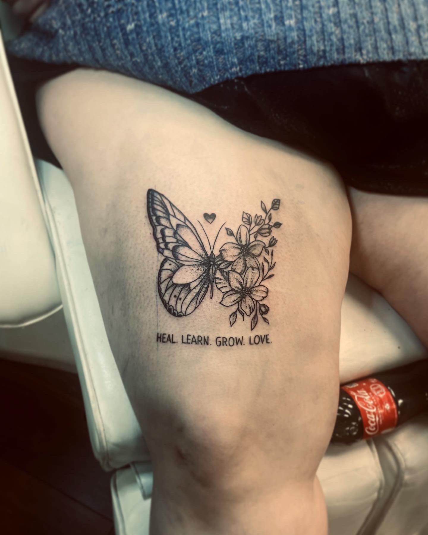 A tattoo on a person's upper thigh featuring a butterfly half-wing and a floral half-wing with text reading 