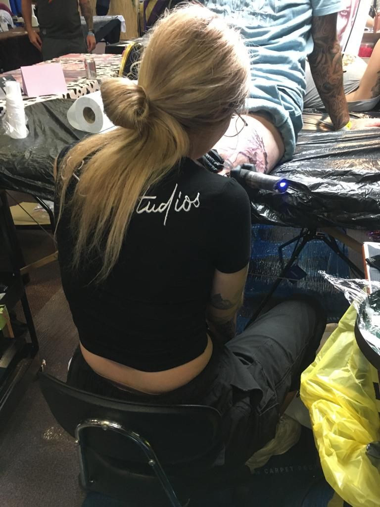 A person with blonde hair in a black shirt tattoos a client's leg inside a studio workspace.