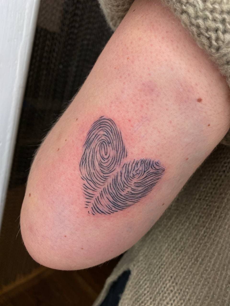 A tattoo on an arm featuring two fingerprints arranged to form a heart shape in black ink.