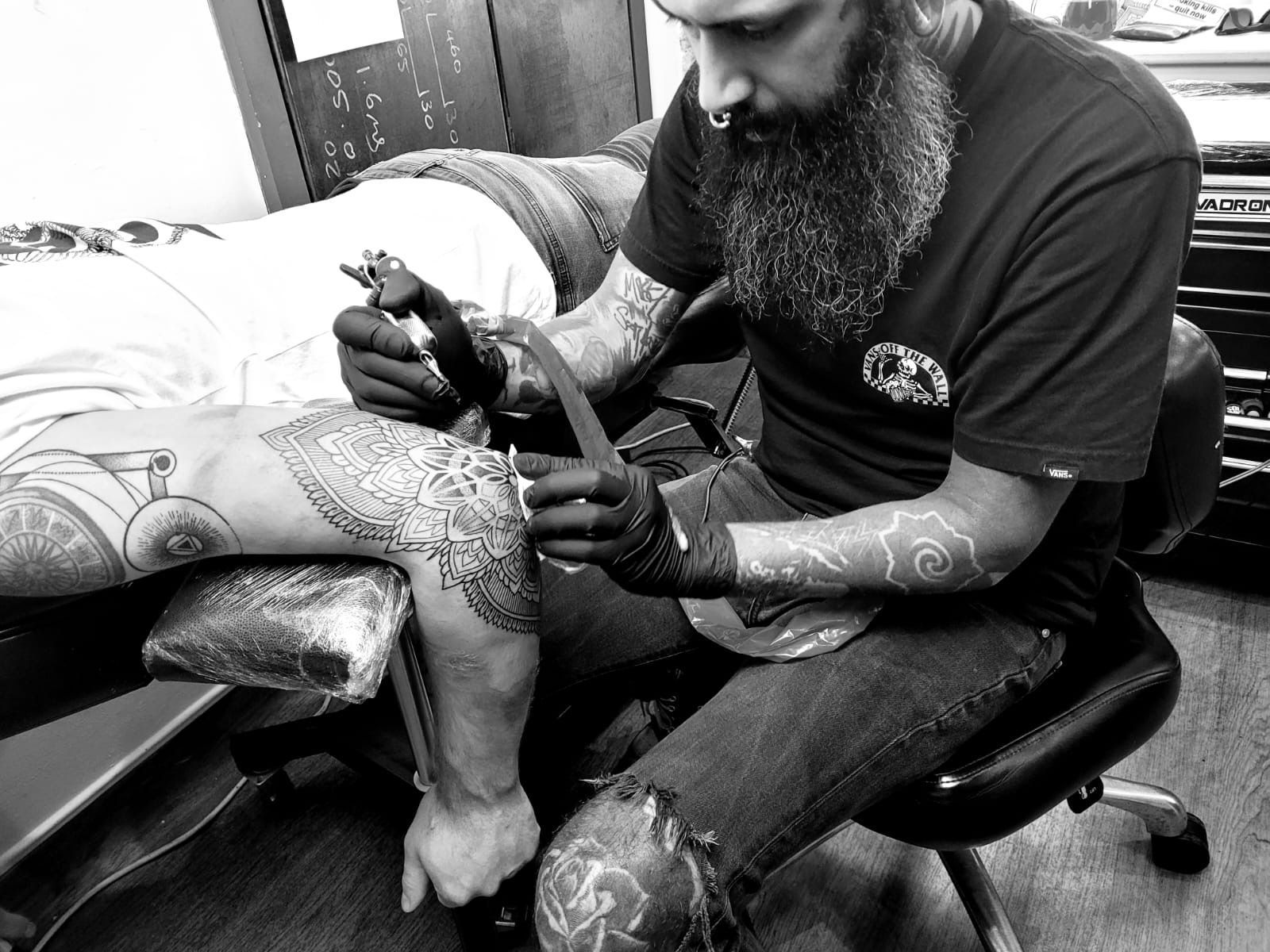 A bearded tattoo artist wearing gloves works on a large, intricate tattoo on a client's arm in a studio.