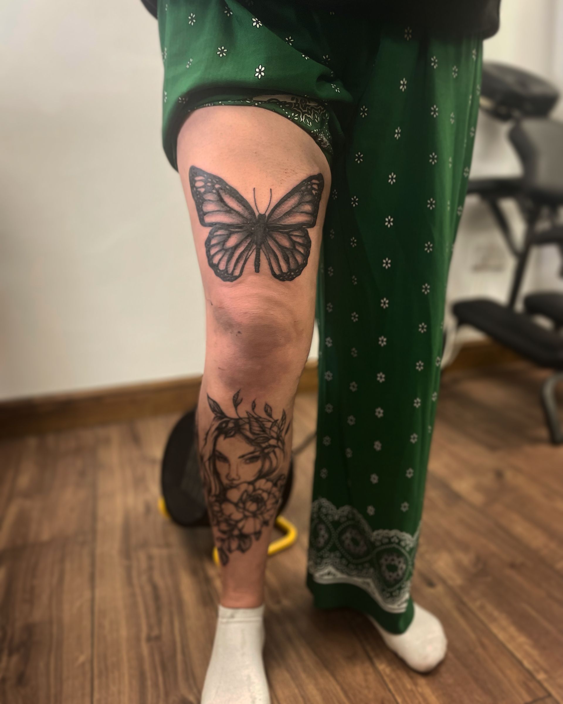 A butterfly tattoo on a thigh and a woman's face surrounded by flowers on the lower leg, seen indoors.