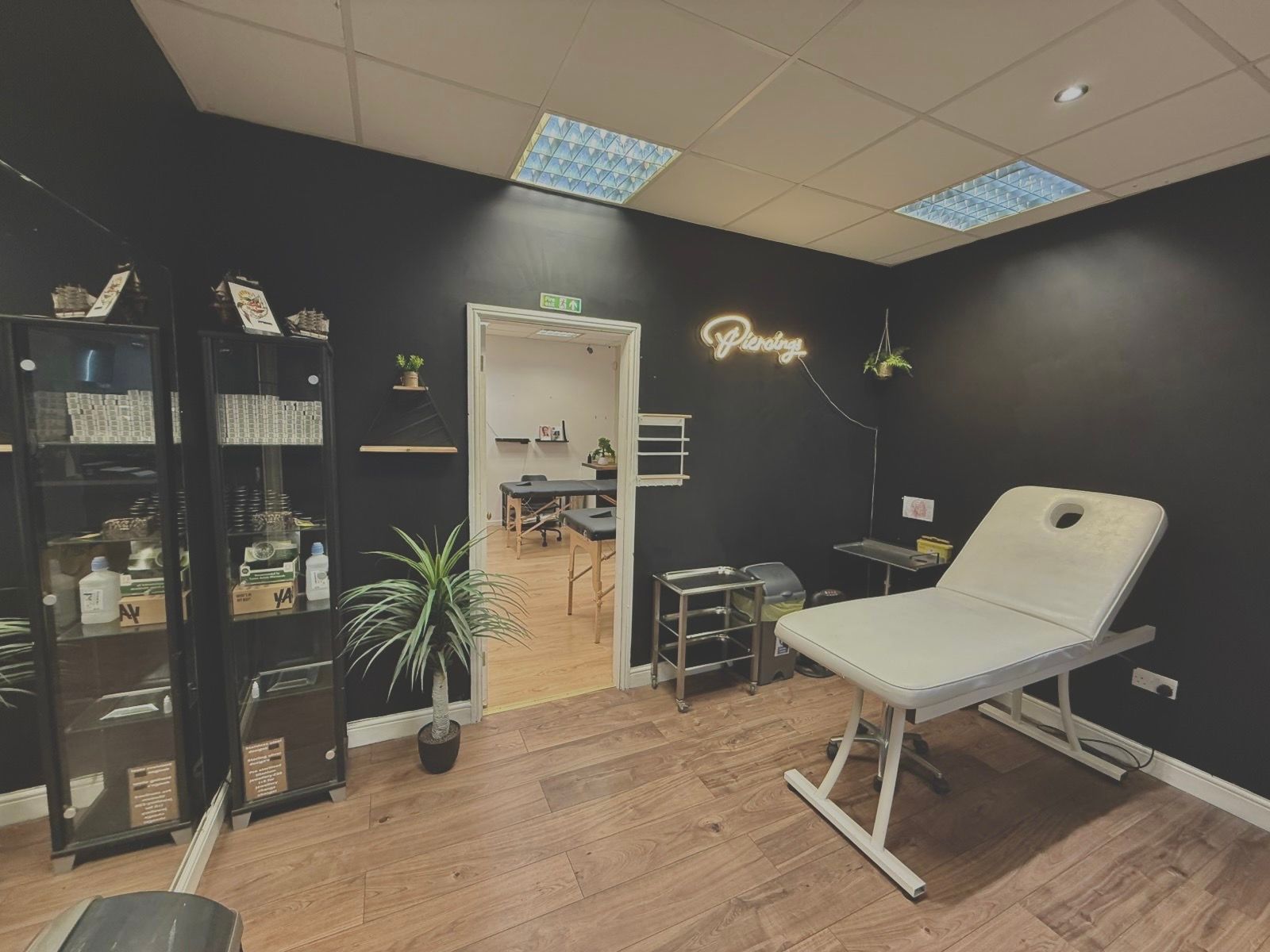 A modern treatment room with dark walls, a white examination bed, glass storage cabinets, a potted plant, and a neon sign.