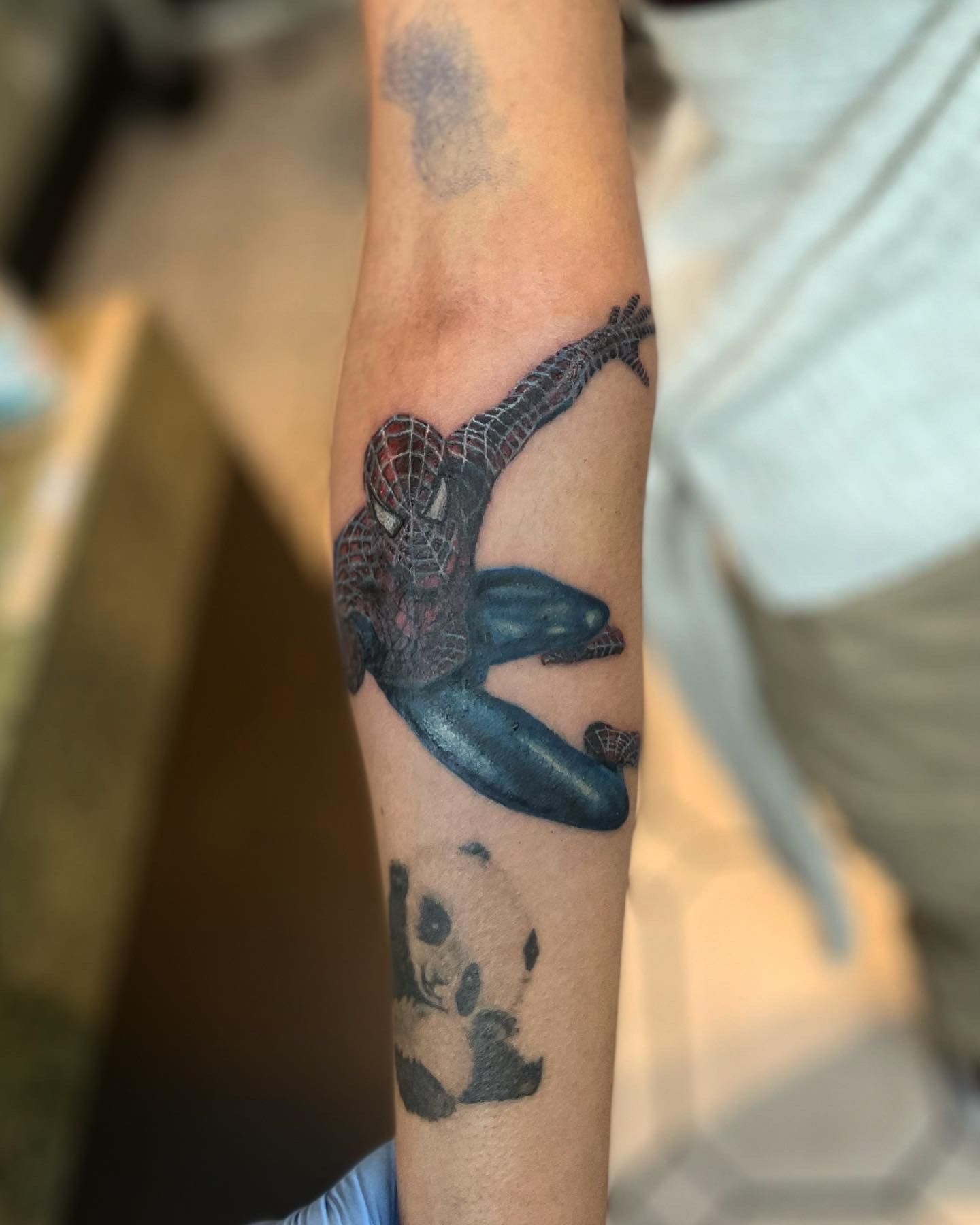 A forearm tattoo of Spider-Man in a dynamic pose above a smaller tattoo of a panda.