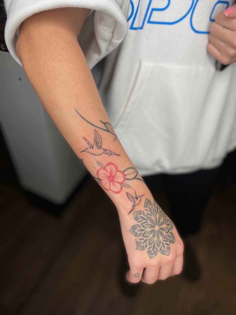 A person's forearm and hand featuring tattoos of a red flower, birds, and a mandala design.