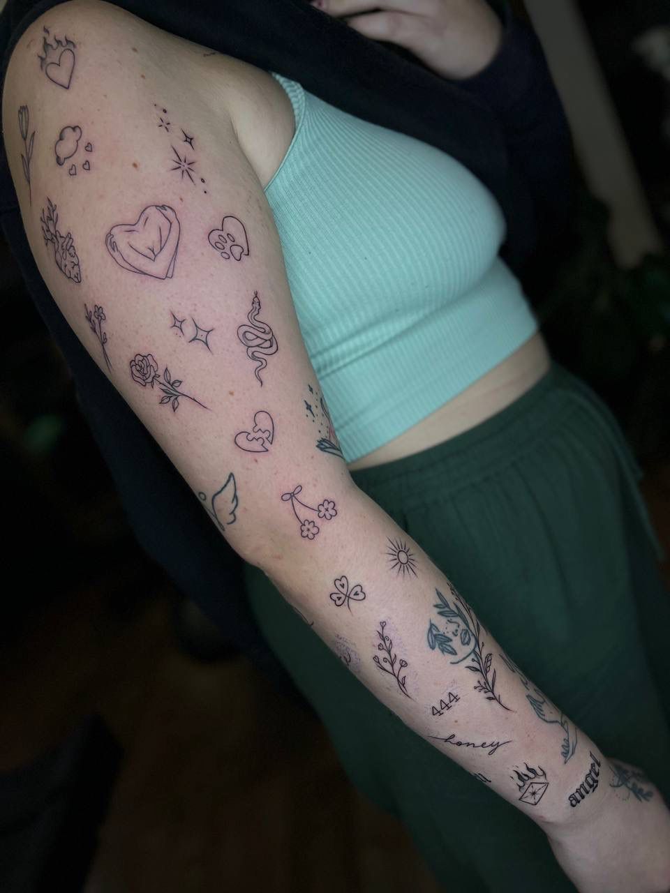 A close-up of an arm covered in multiple small, fine-line tattoos, including hearts, stars, flowers, and a snake.
