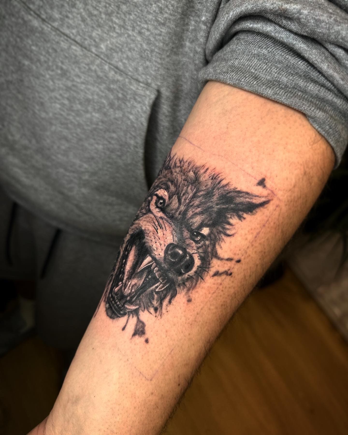 A forearm tattoo of a snarling wolf head with textured black ink detailing against grey clothing.