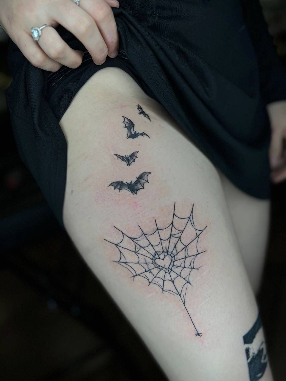 A black ink tattoo on a thigh featuring four flying bats ascending toward a heart-shaped spider web.