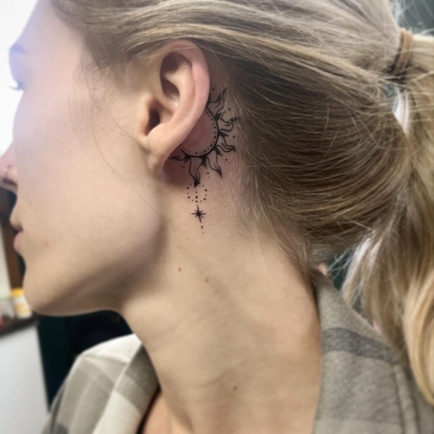 A detailed, fine-line tattoo of a sun with an attached star charm, located directly behind a person's ear.