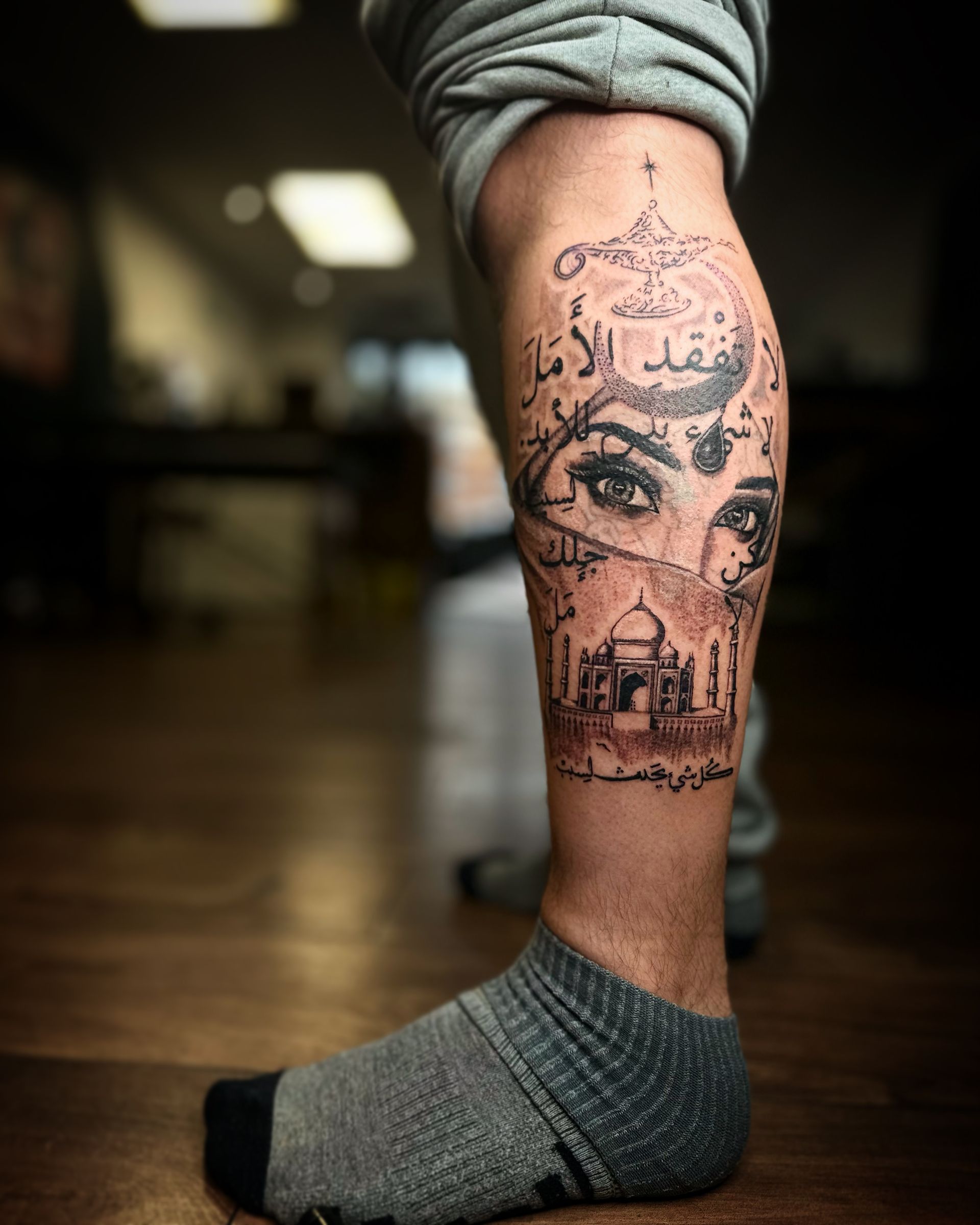 A tattoo on a lower leg featuring a woman's eyes, a dome structure, a lamp, and Arabic calligraphy.