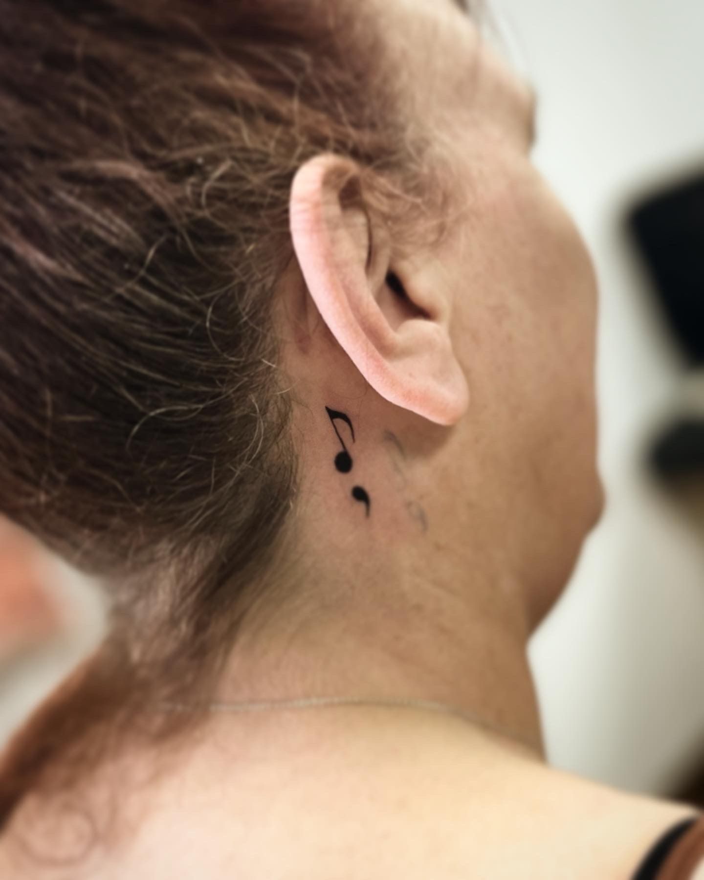 A small black tattoo of a musical note combined with a semicolon behind a person's ear.