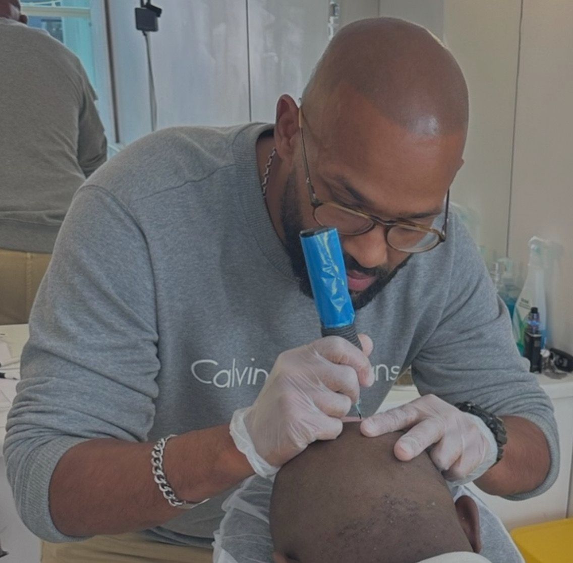 A person in a gray sweatshirt performing a scalp micropigmentation procedure on a client's head with a tattoo device.
