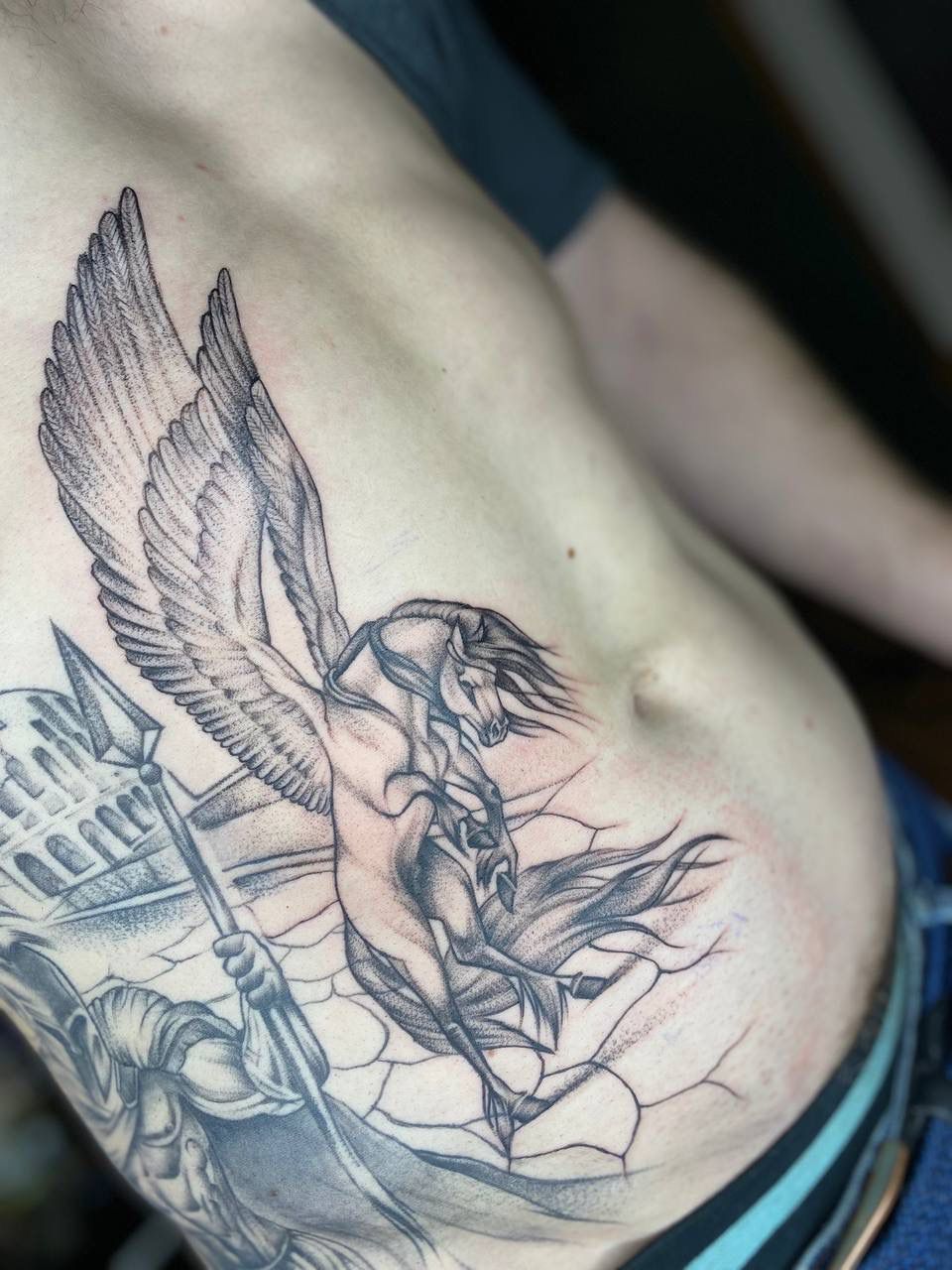 Black and grey tattoo on a torso depicting a winged horse soaring above a classical building and a spear-wielding figure.