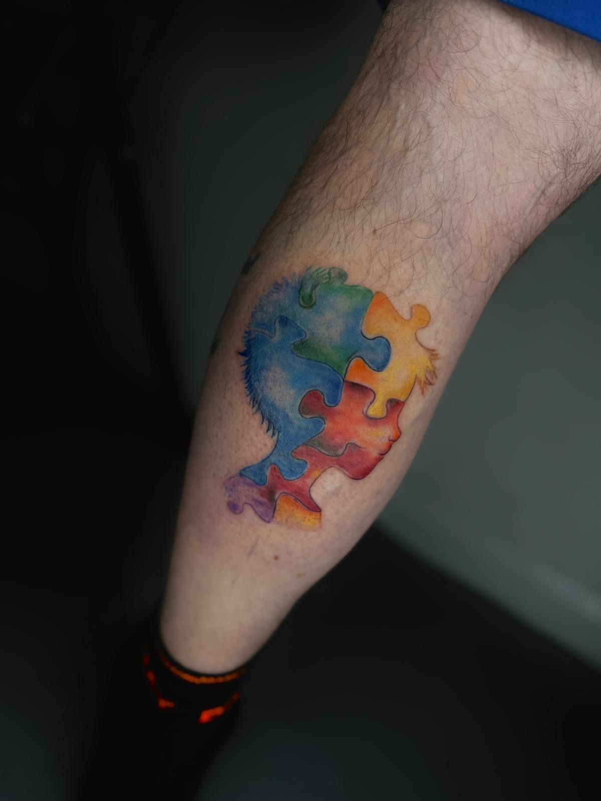 A tattoo on a calf depicting a human head silhouette formed by interconnected, multi-colored watercolor puzzle pieces.