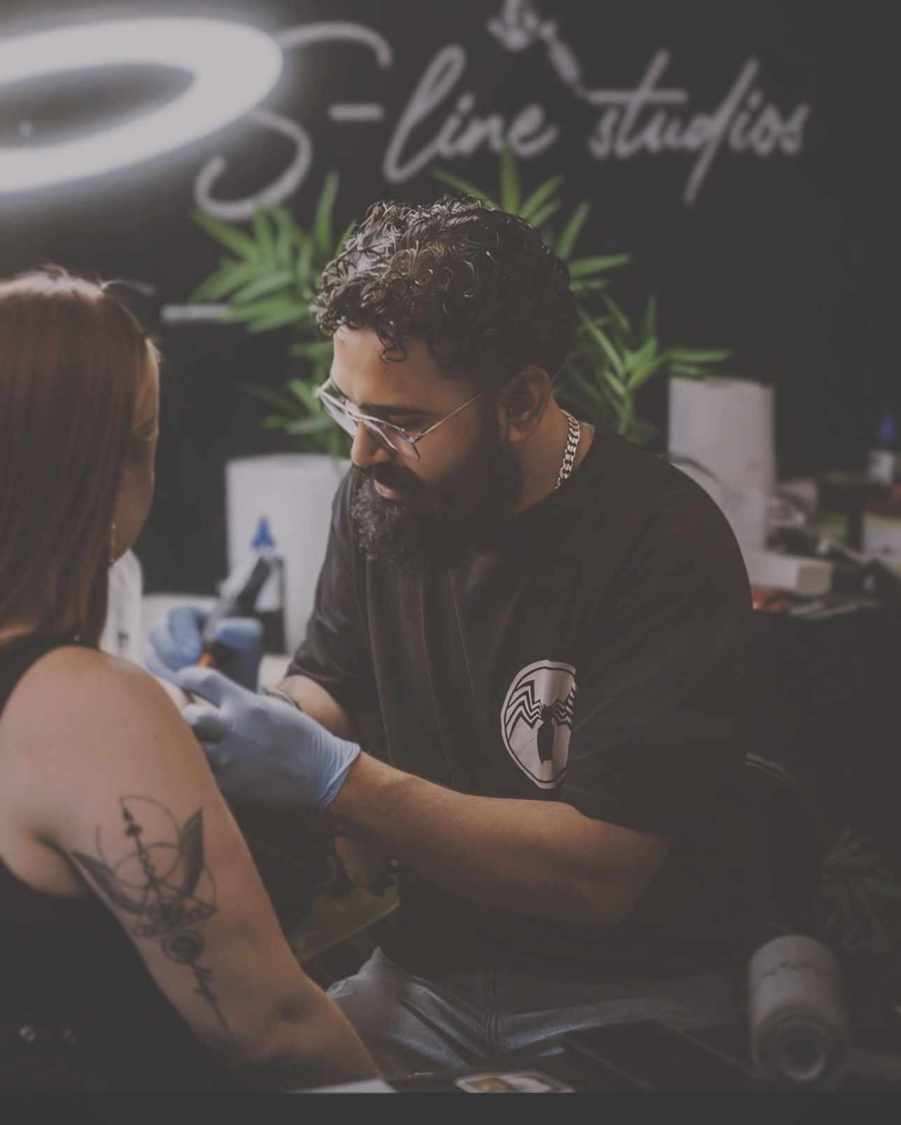 A tattoo artist wearing gloves and glasses works on a client’s shoulder inside S-line studios under bright ring lighting.