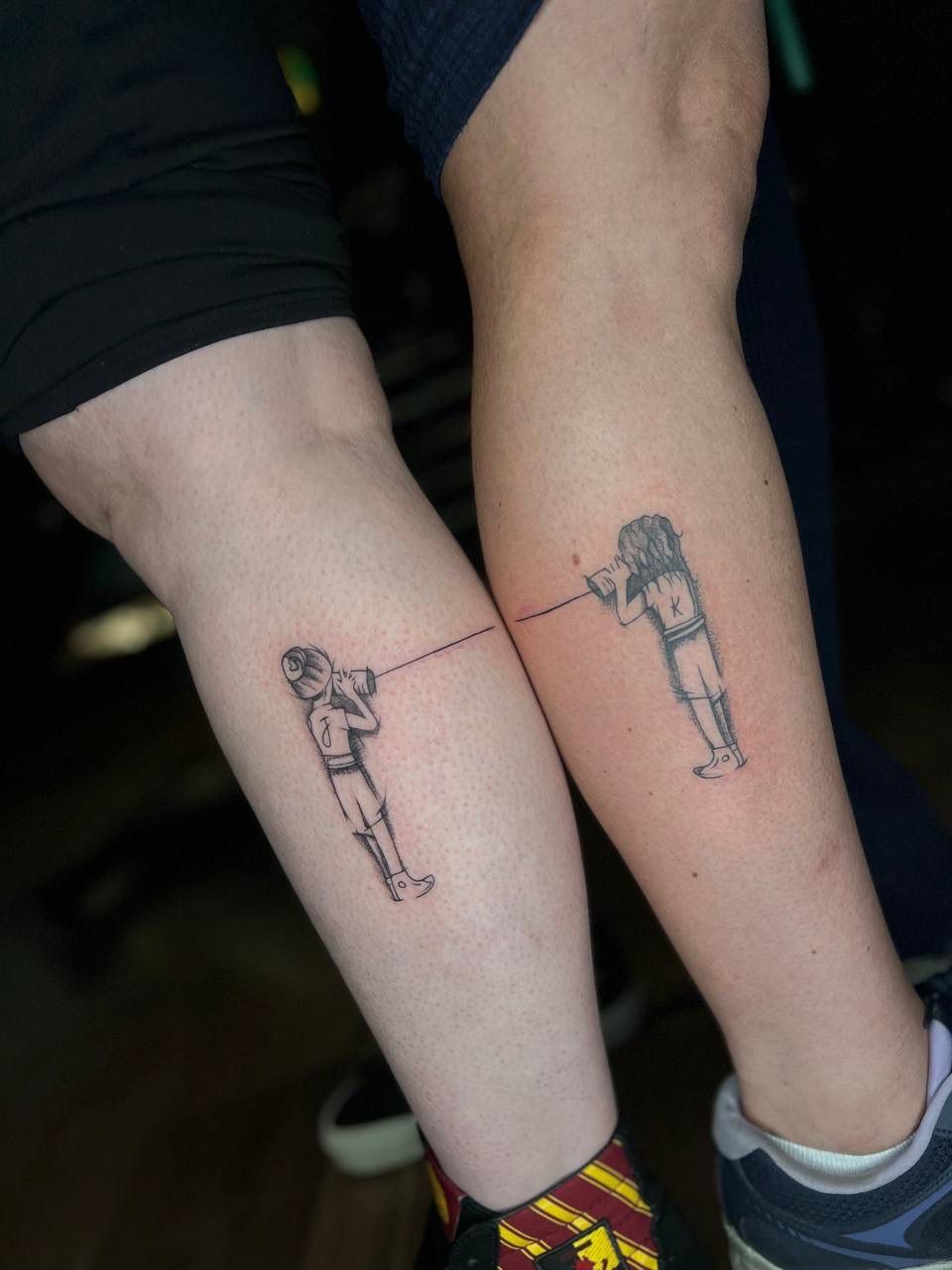 A matching tattoo on two legs showing two figures connected by a string-and-cup telephone, each holding a cup to their ear.
