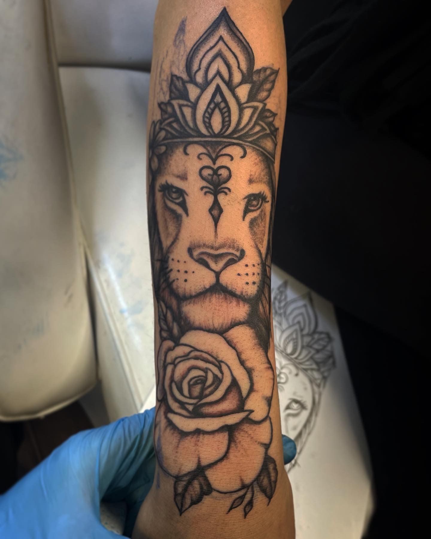 A forearm tattoo of a lion adorned with a decorative crown and a floral design below it, rendered in black and grey ink.