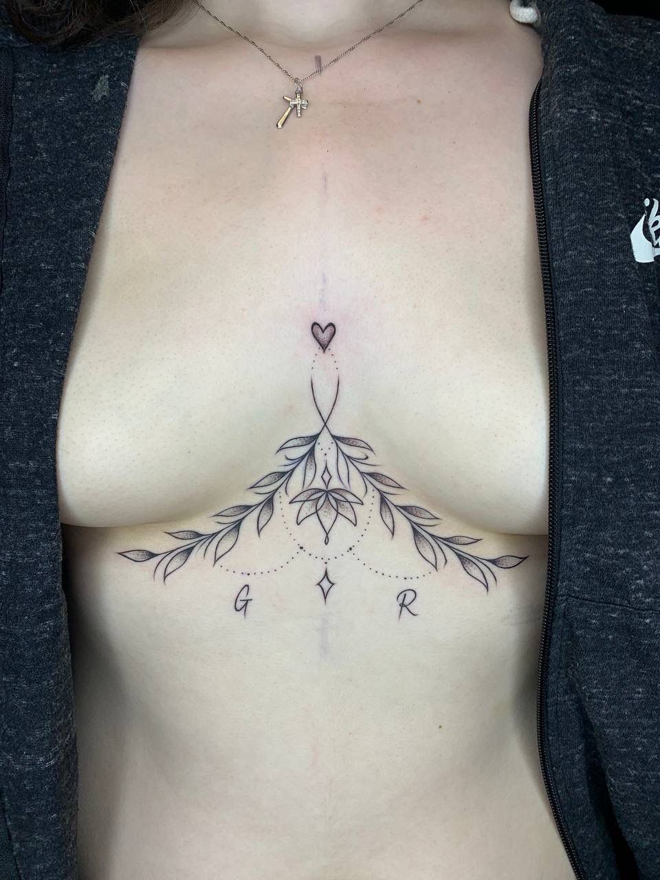 A sternum tattoo featuring a small heart, botanical branches, a central flower, and the letters G and R.