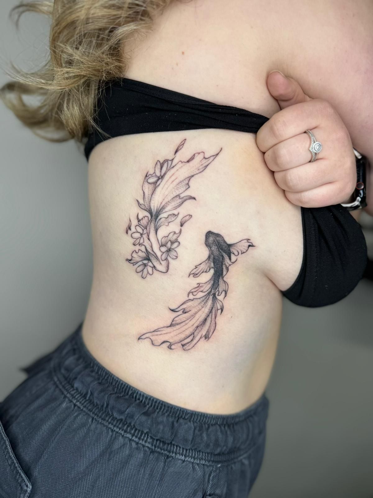 A side-profile view of a ribcage tattoo featuring two stylized betta fish with flowing fins and cherry blossom accents.