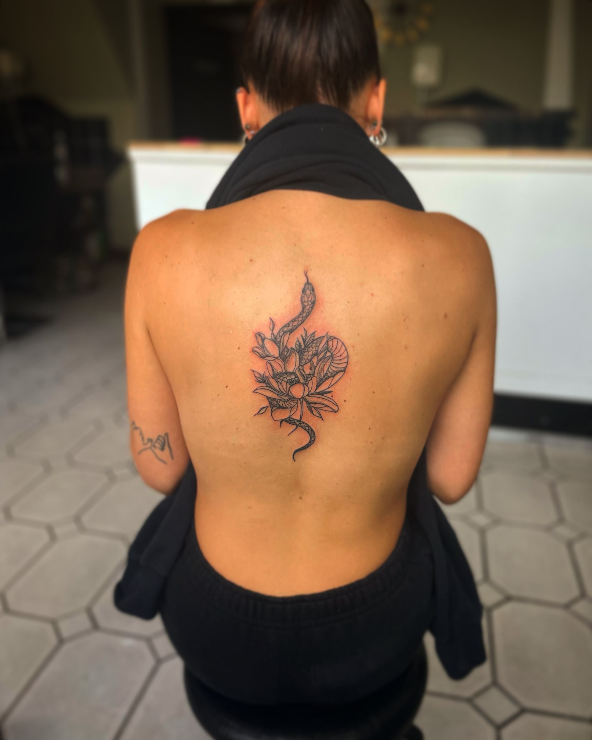 A person seen from behind, wearing a backless top, showcasing a fine-line tattoo of a snake coiled around a flower.
