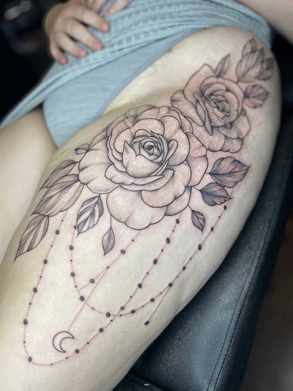 Thigh tattoo of two roses with leaves and hanging beaded chains featuring a crescent moon charm in black ink.