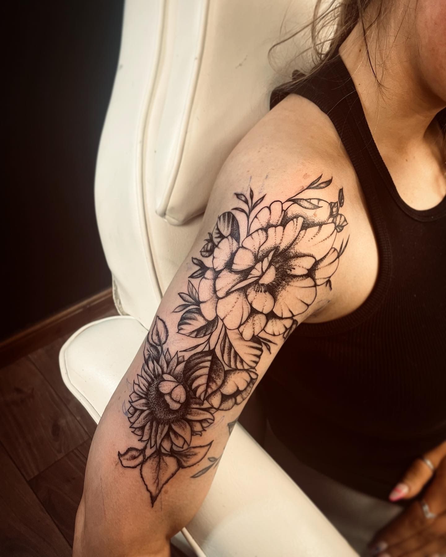 A close-up view of a black-ink floral tattoo featuring large peonies and a sunflower on the side of a person's upper arm.