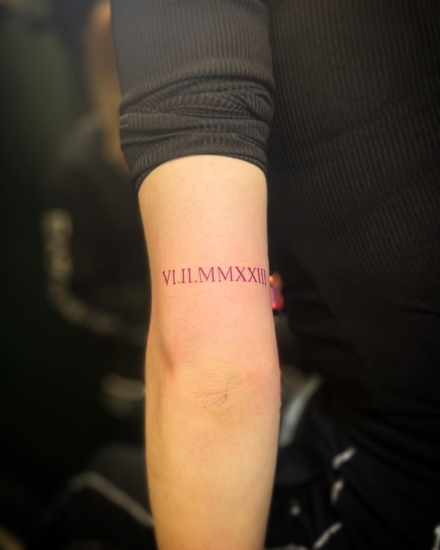 A close-up of a forearm featuring a minimalist red tattoo with Roman numerals reading 
