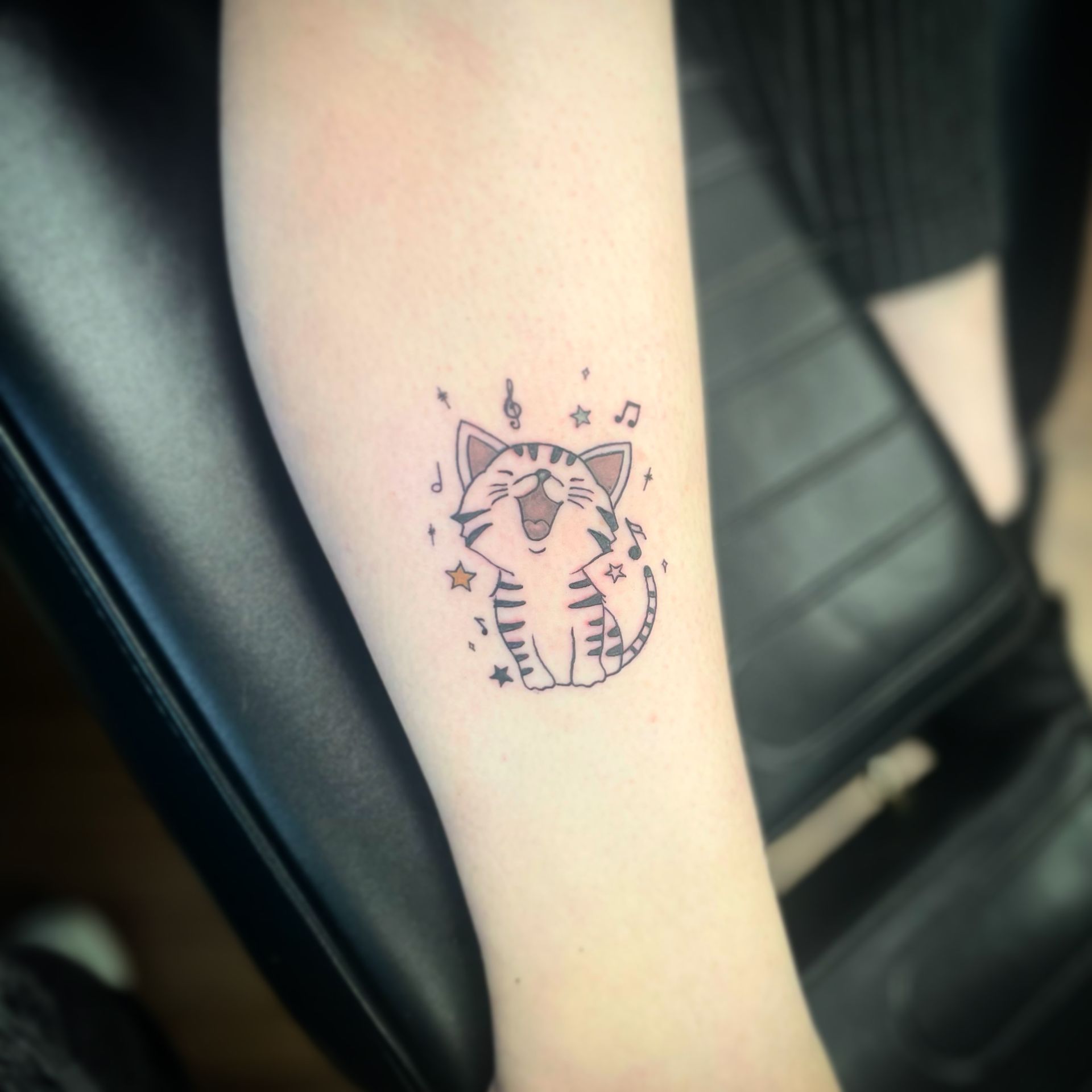 A small, fine-line tattoo on an arm featuring a cute, singing tabby kitten surrounded by musical notes and stars.
