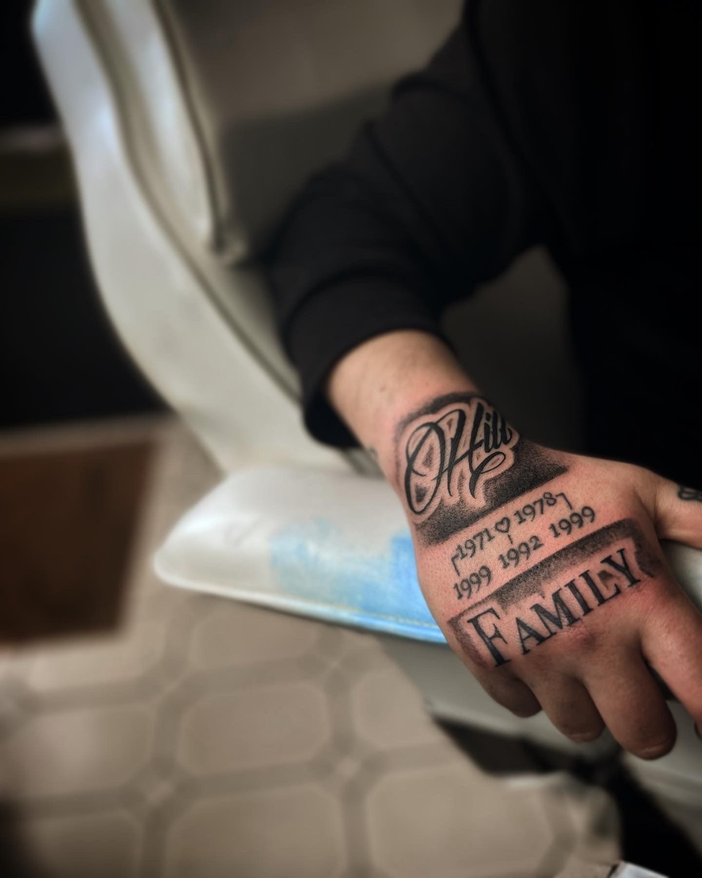 A close-up of a hand with black and grey tattoos reading 