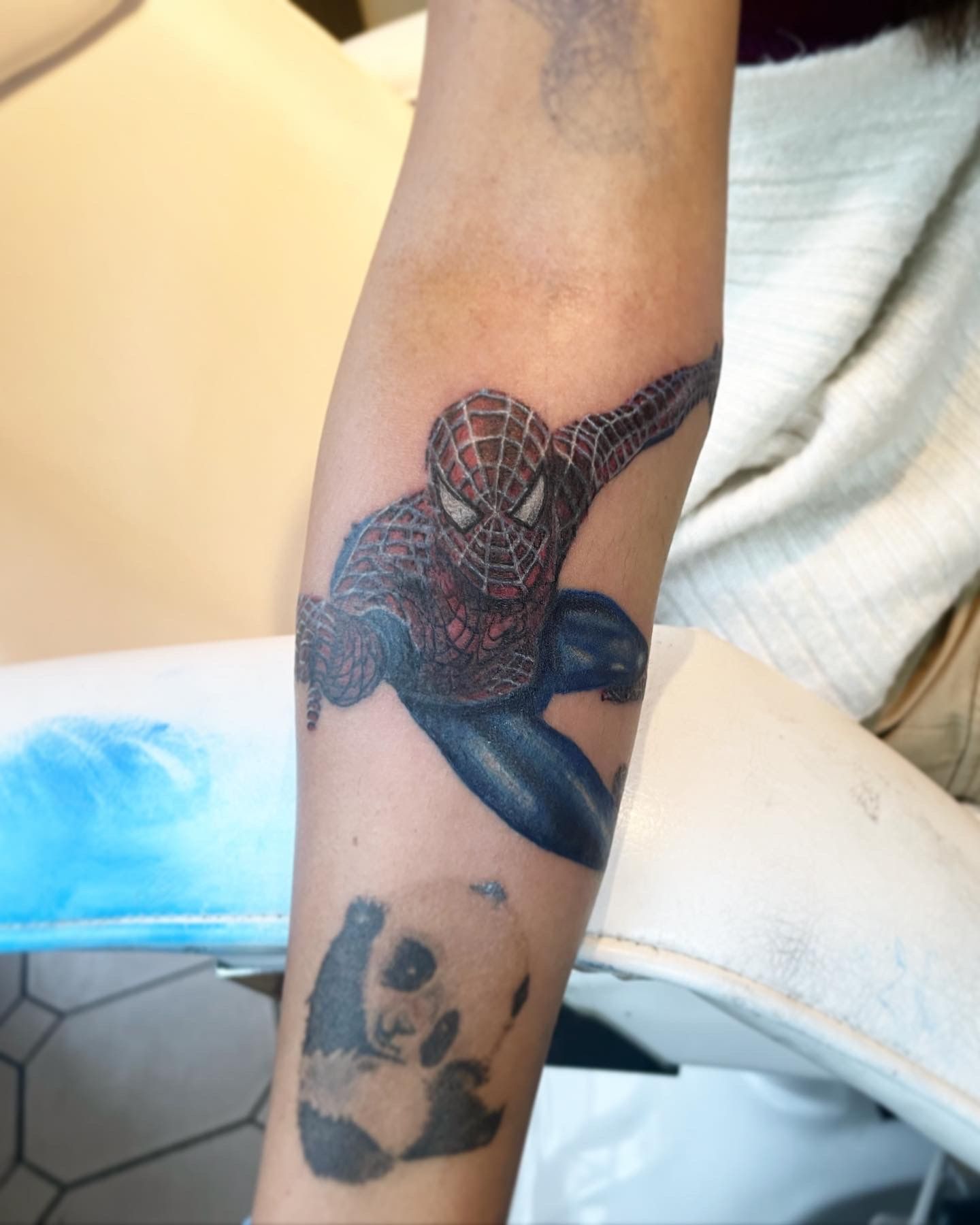 A forearm tattoo featuring a dynamic, textured Spider-Man above a small, shaded panda portrait.