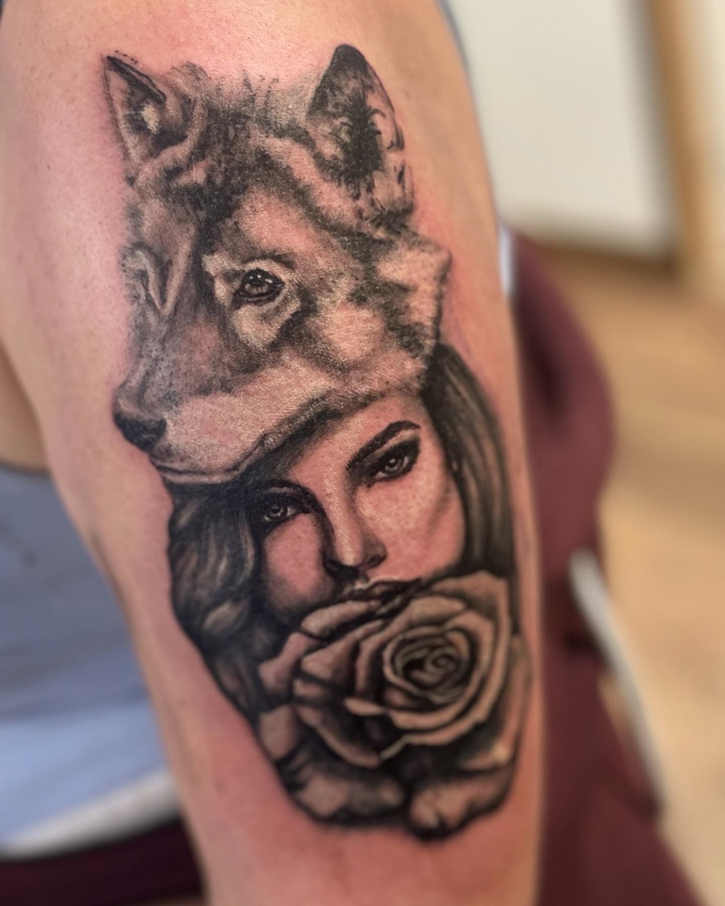 A black-and-grey tattoo on skin showing a person's face wearing a wolf pelt headdress, with a rose held to their mouth.