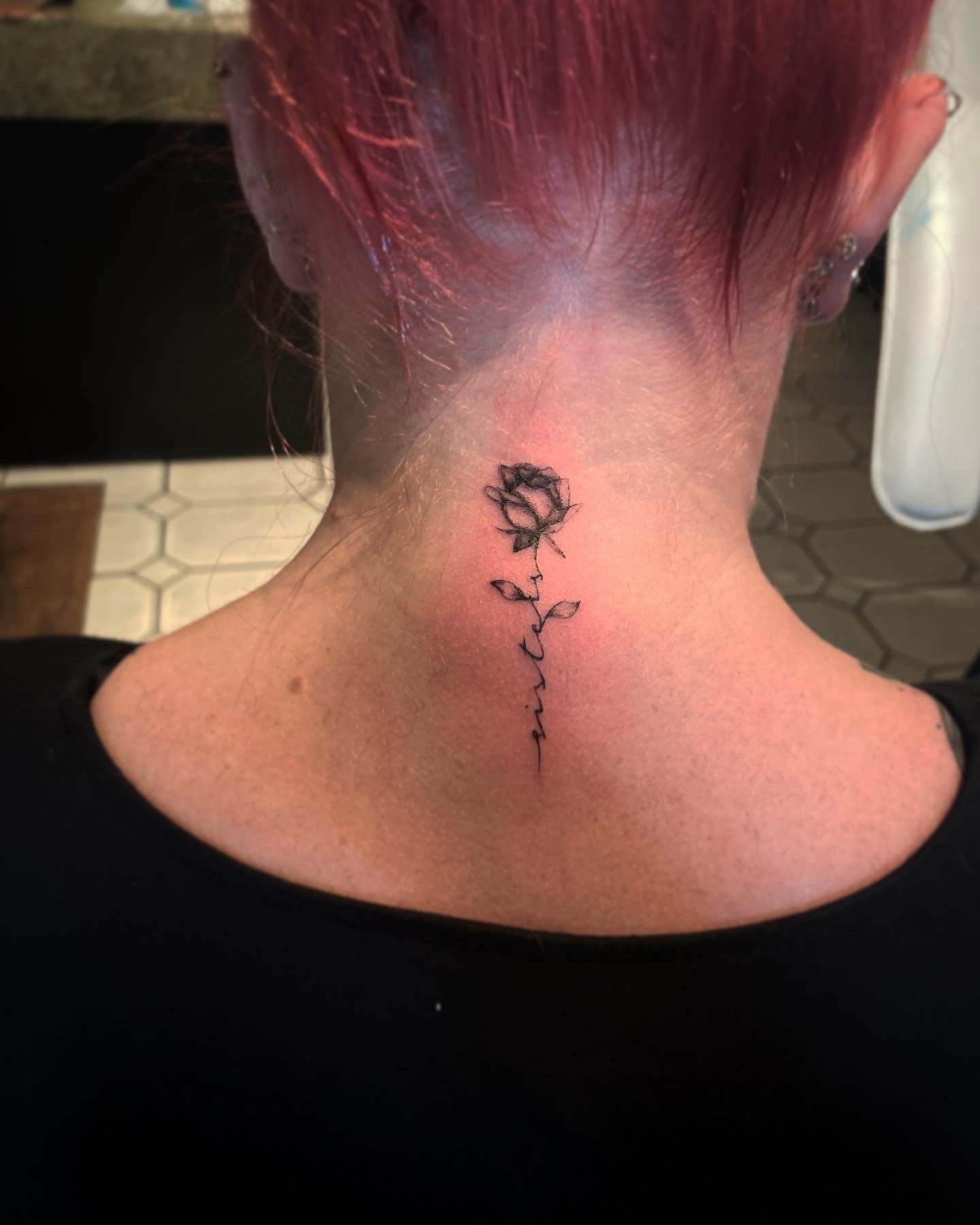 A delicate, sketch-style rose tattoo on the back of a neck, with a thin, flowing stem extending downward.