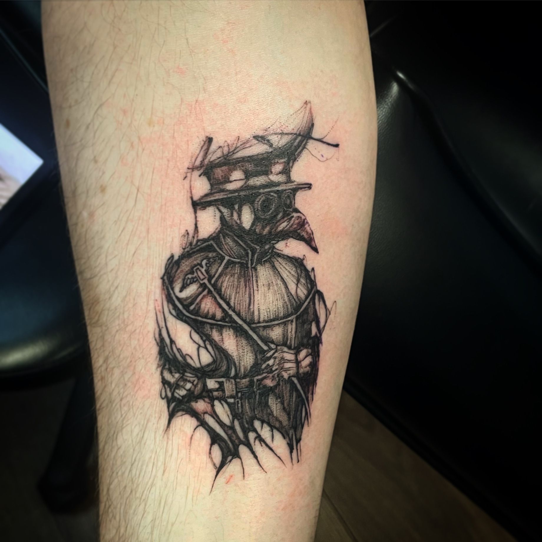 A black and white tattoo on a person's forearm featuring a stylized plague doctor in a sketchy, ink-washed art style.