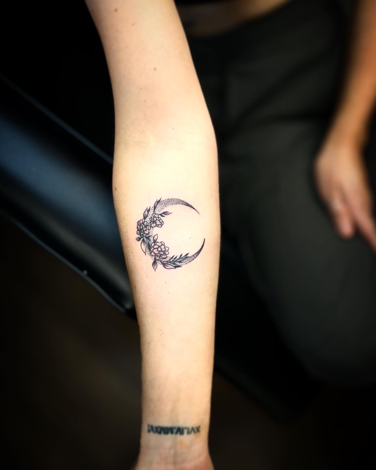 A person's inner forearm featuring a small crescent moon tattoo made of intricate black-ink floral patterns.