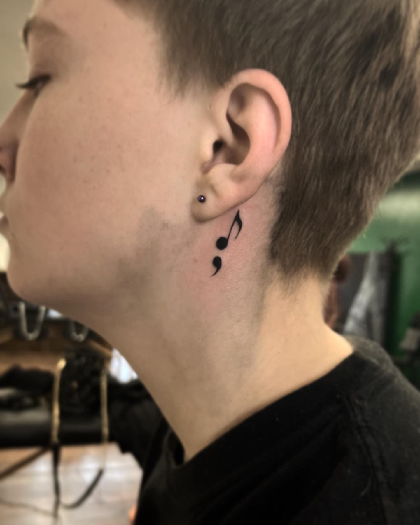 A person with a black ink tattoo behind their ear combining a musical note and a semicolon.