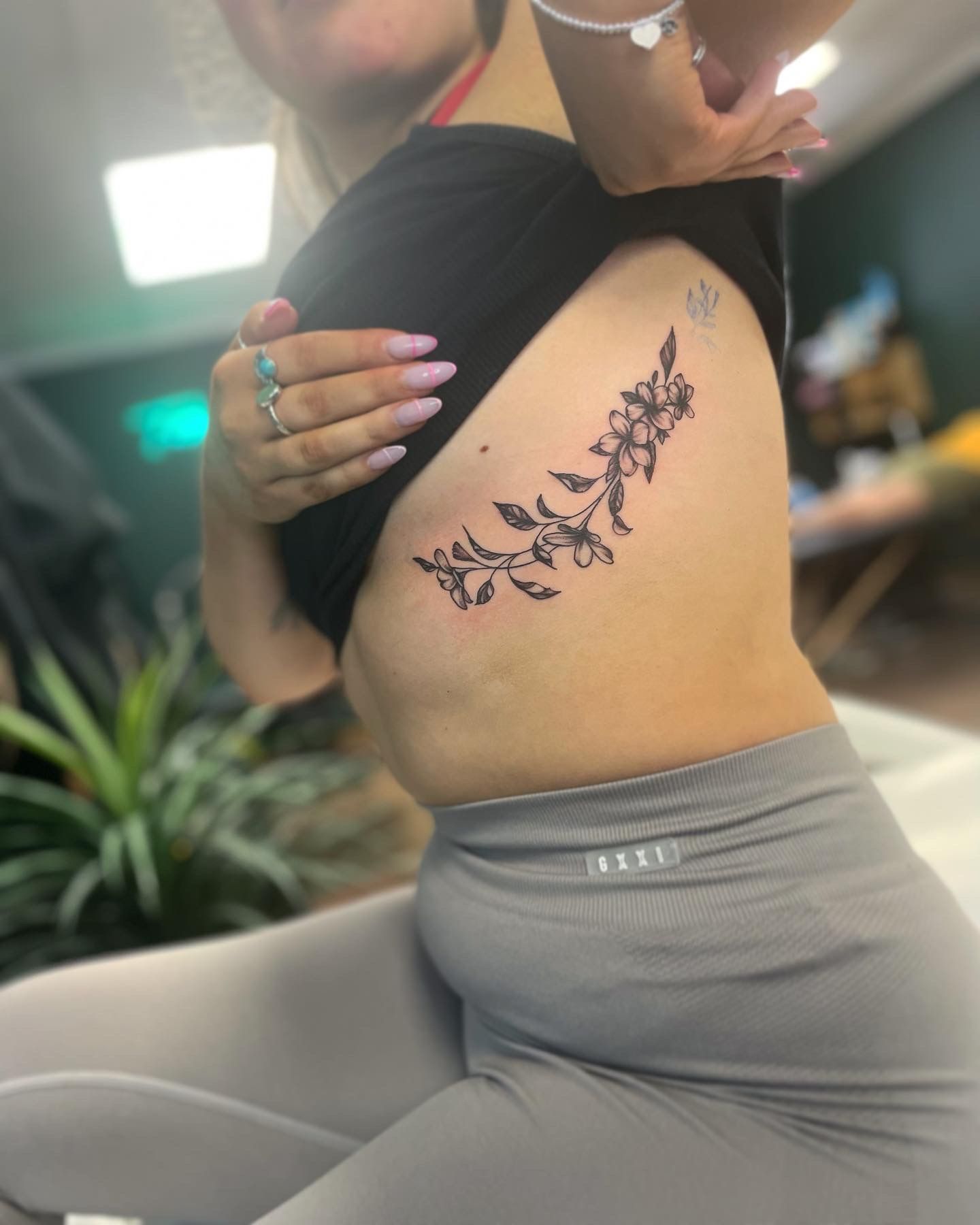 A person shows a delicate floral vine tattoo on their ribcage, featuring black linework with small blossoms and leaves.