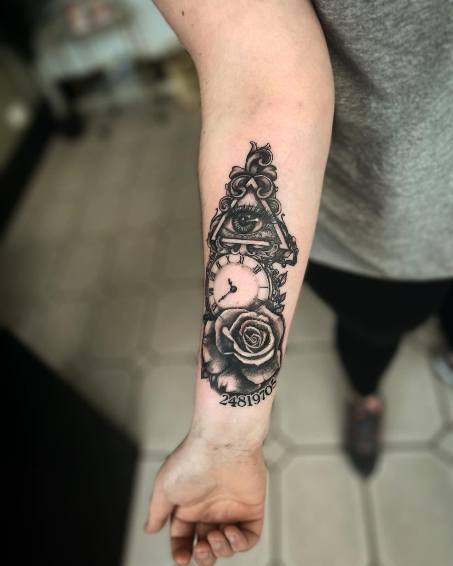 A black-and-grey forearm tattoo featuring an All-Seeing Eye above a clock, a rose, and a numerical date.