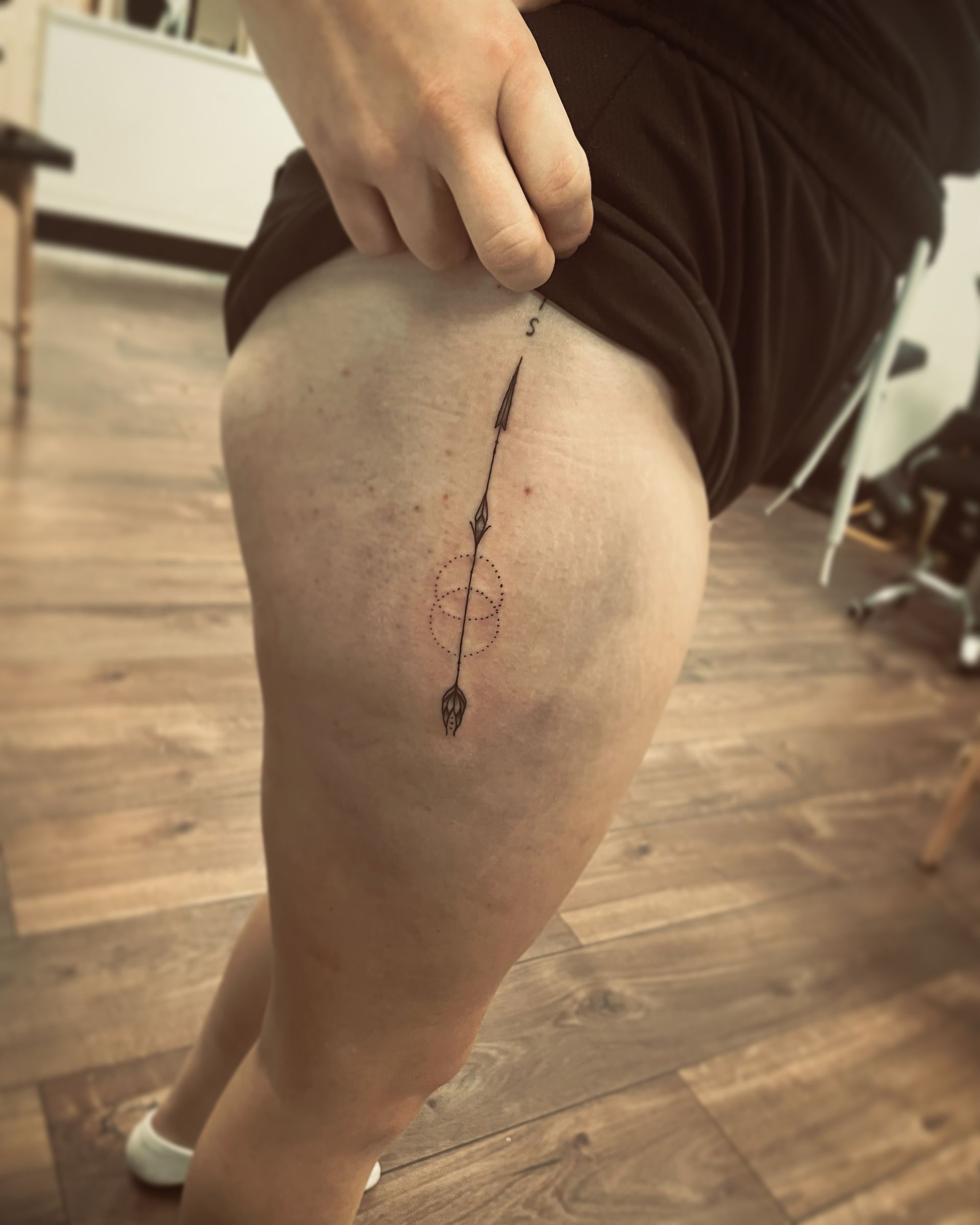 A person holds up the edge of their garment to reveal a thin, vertical fine-line tattoo on their upper thigh.