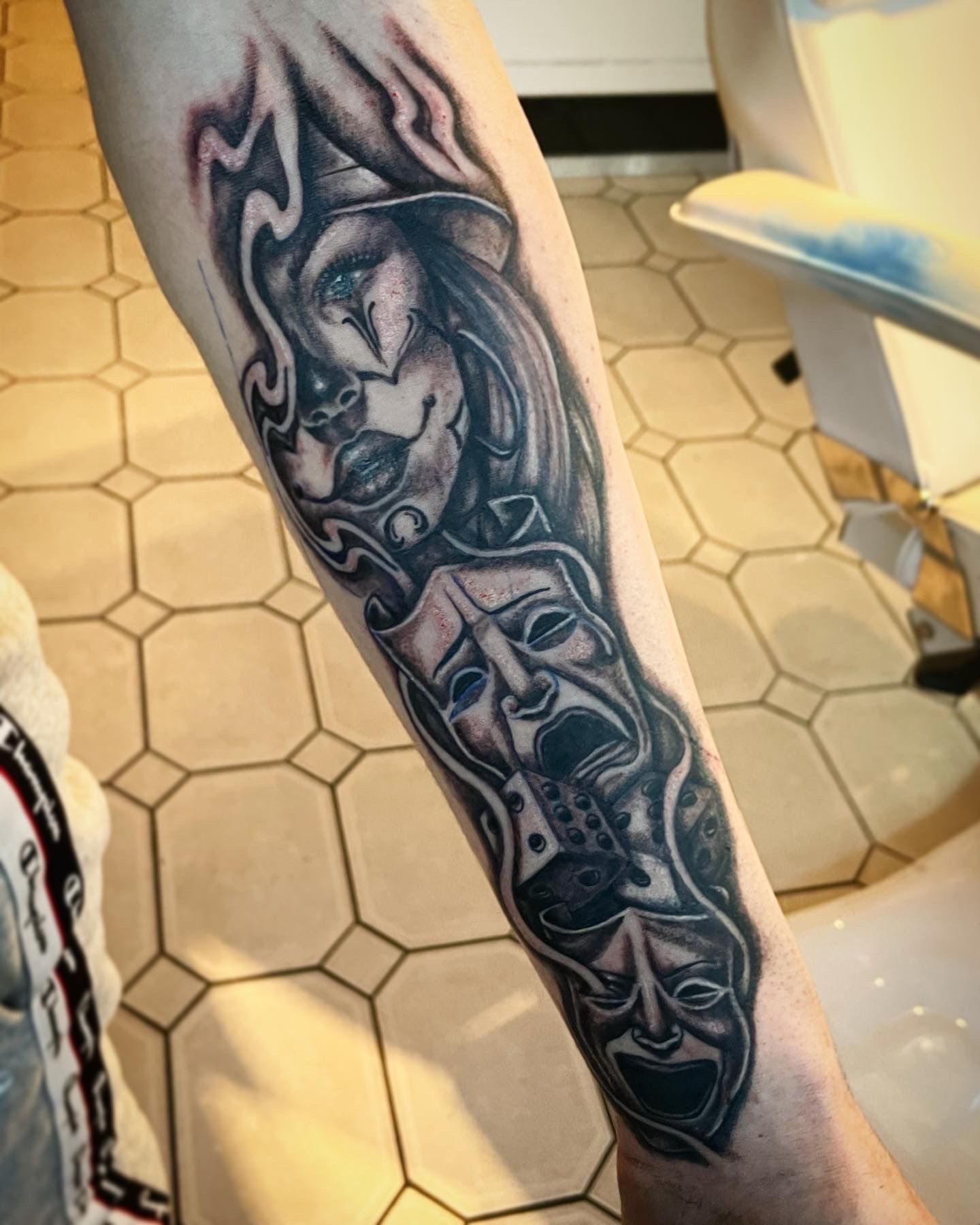 Forearm tattoo featuring a clown, two theatrical tragedy masks, and a die, rendered in detailed black-and-grey shading.