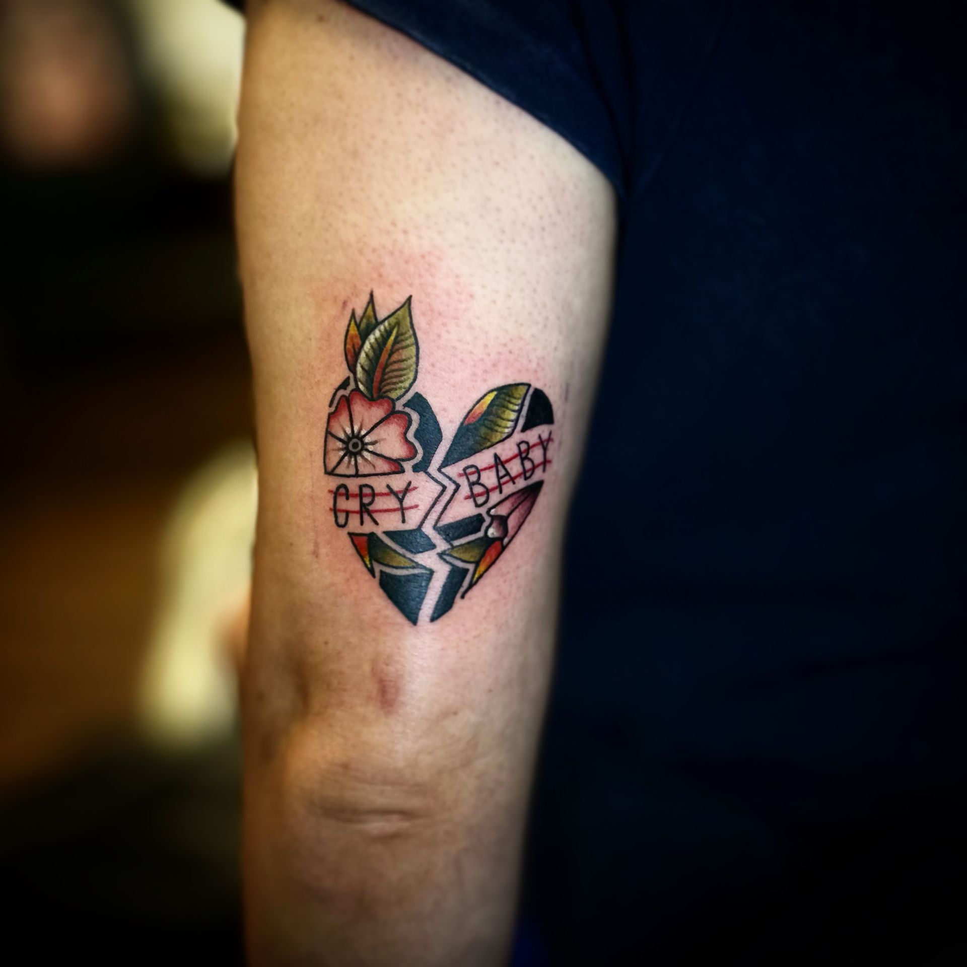 A broken heart tattoo on an arm, featuring green leaves, a pink flower, and the words 