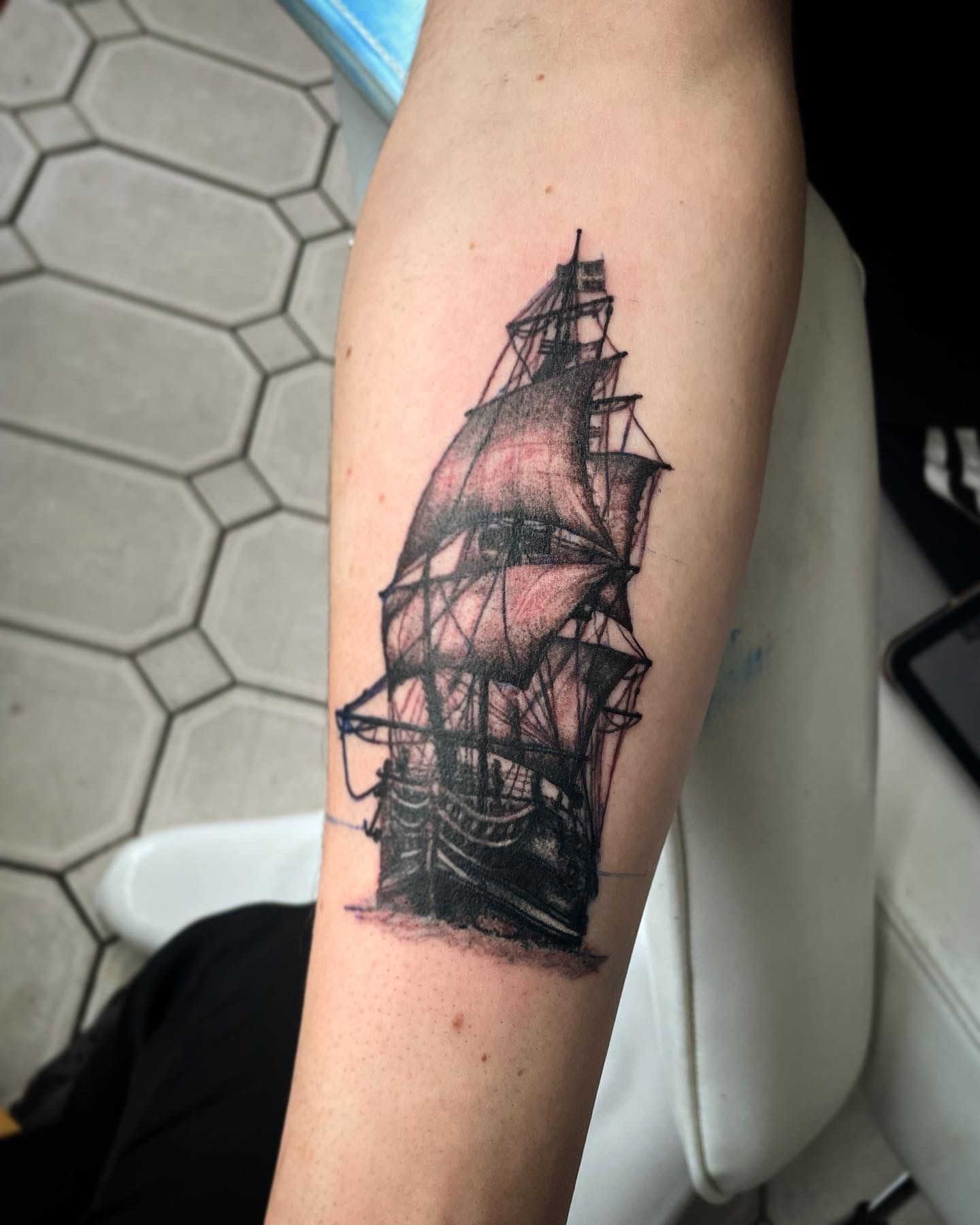 A black and grey, fine-line tattoo of a tall ship with full sails on a person's forearm.