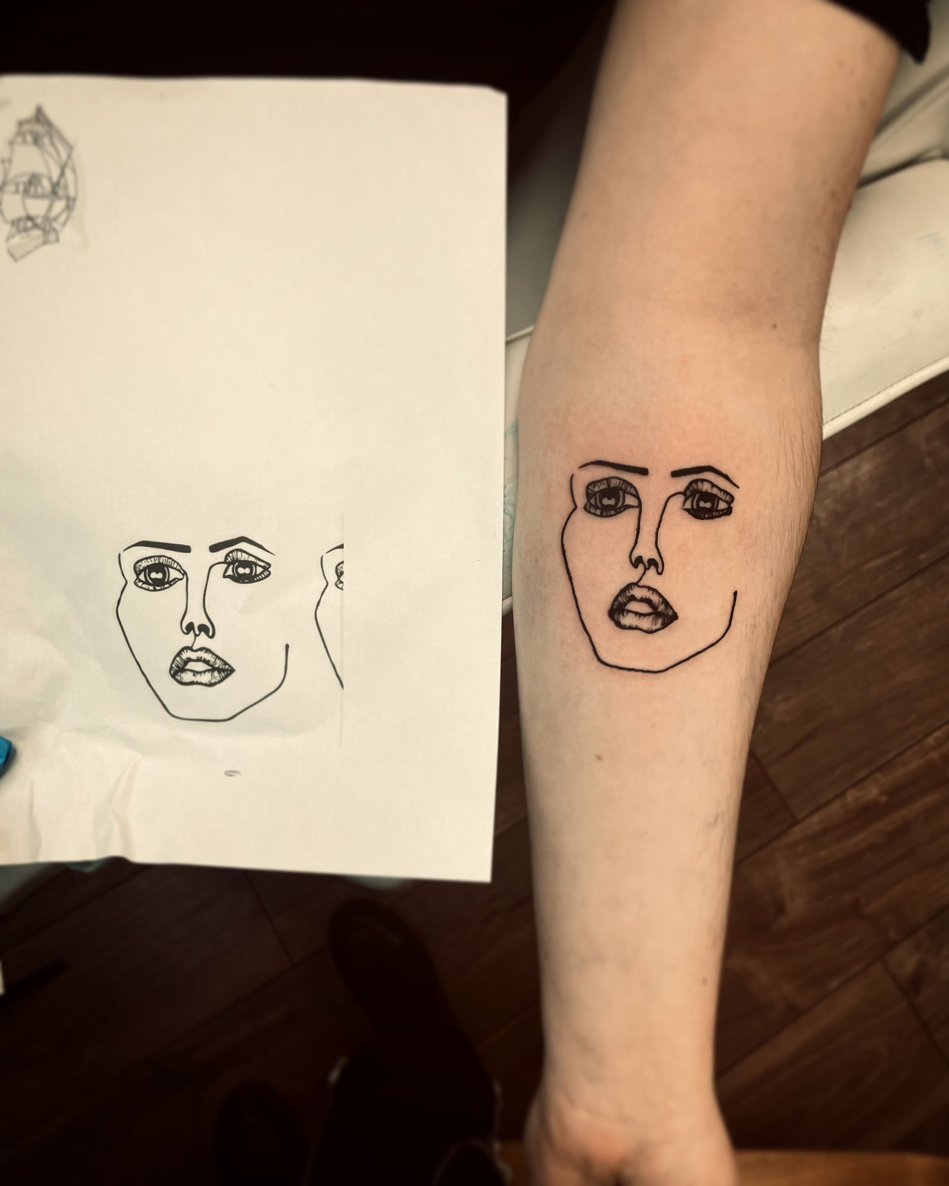 A minimalist line-art drawing of a face on paper held next to an identical tattoo on a person's forearm.