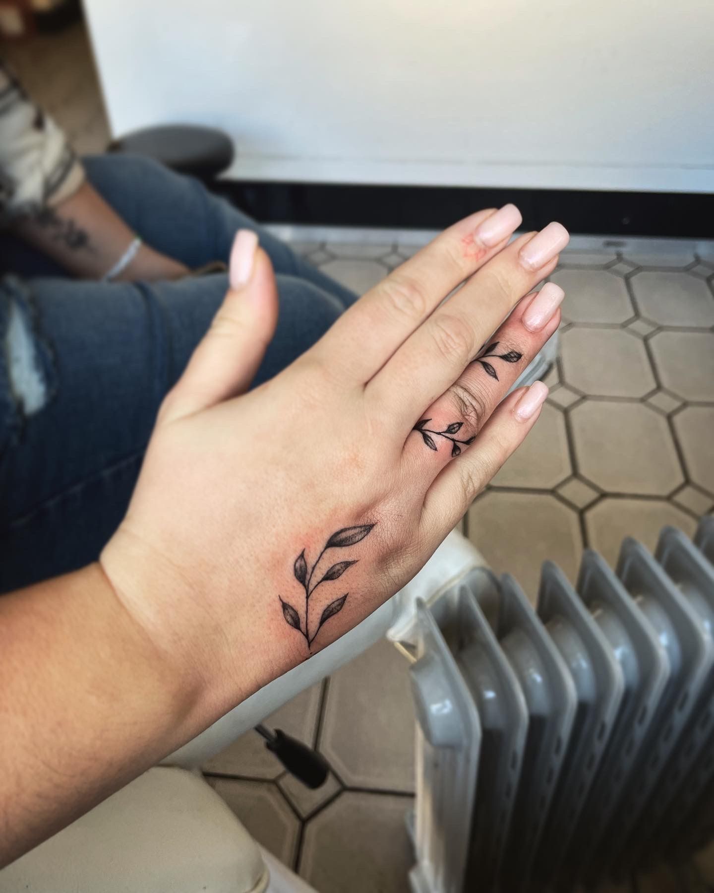 A hand with two delicate black-ink leaf tattoos, one on the back of the hand and one along the middle finger.