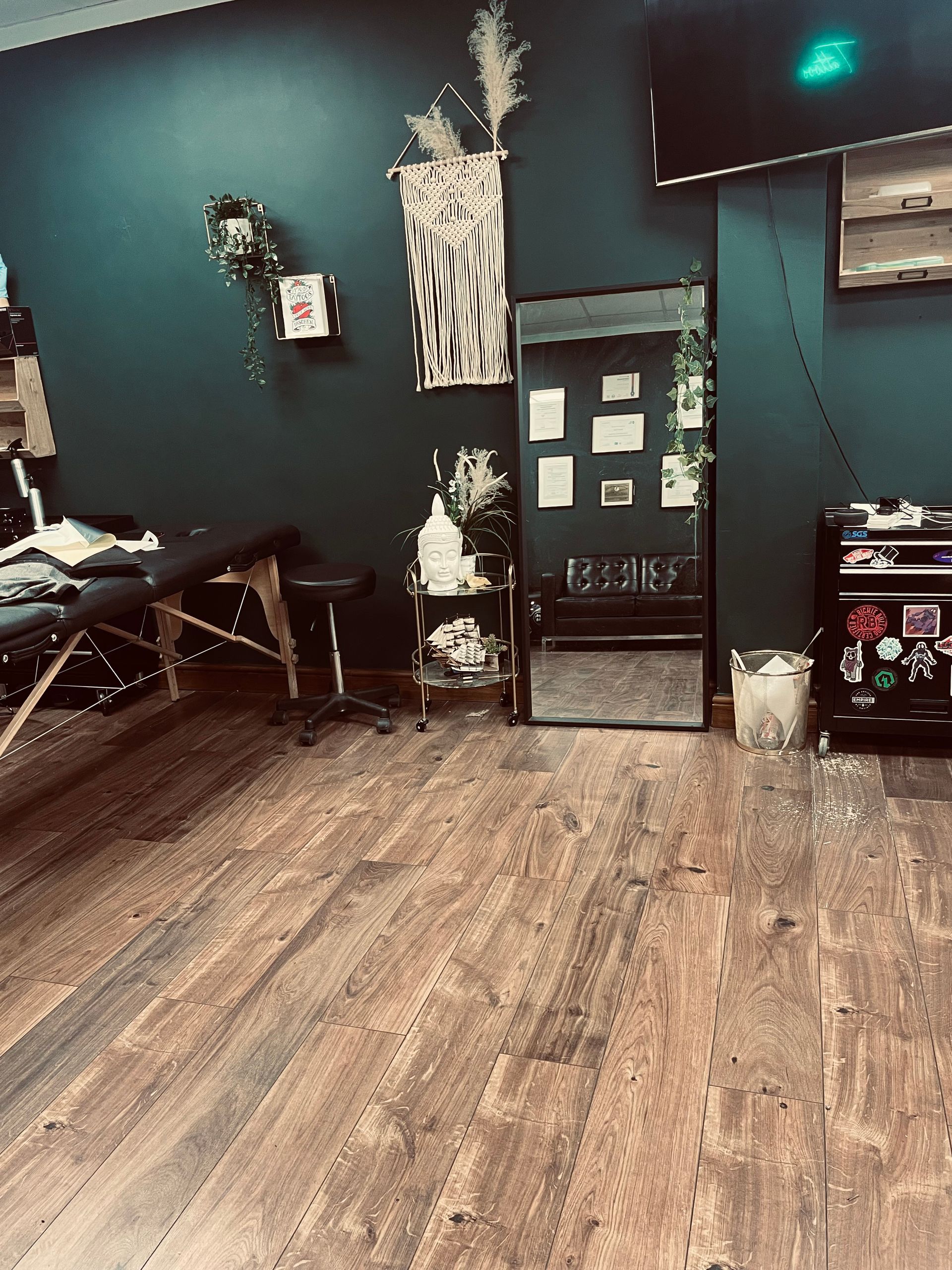 A tattoo studio with dark teal walls, a treatment table, a mirror, wooden floors, and various decor.