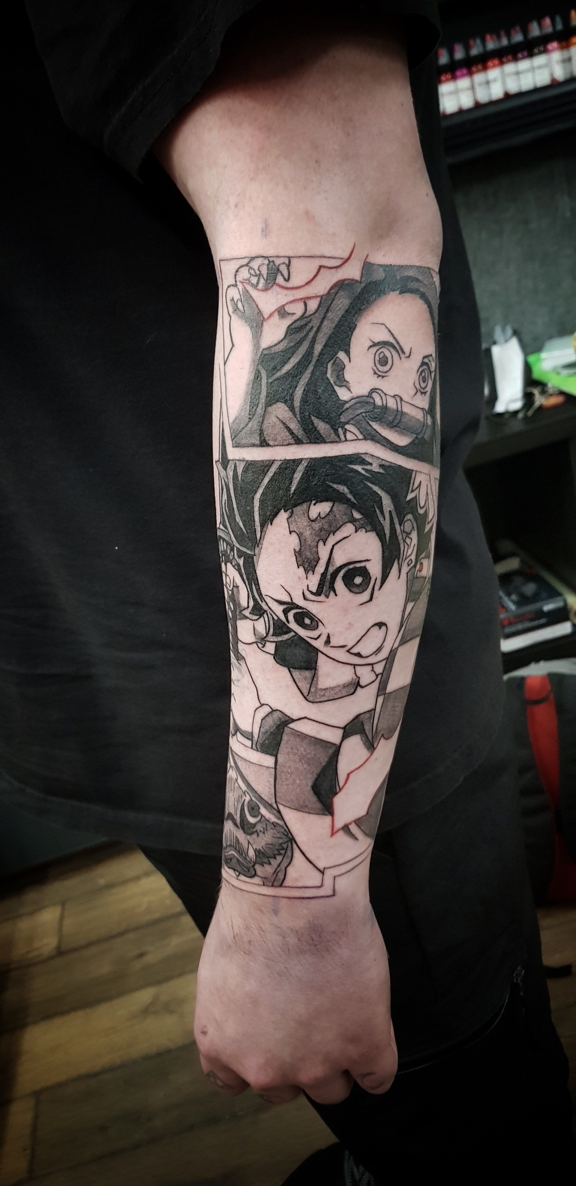 A forearm tattoo featuring manga-style illustrations of Nezuko and Tanjiro from Demon Slayer in black and gray ink.