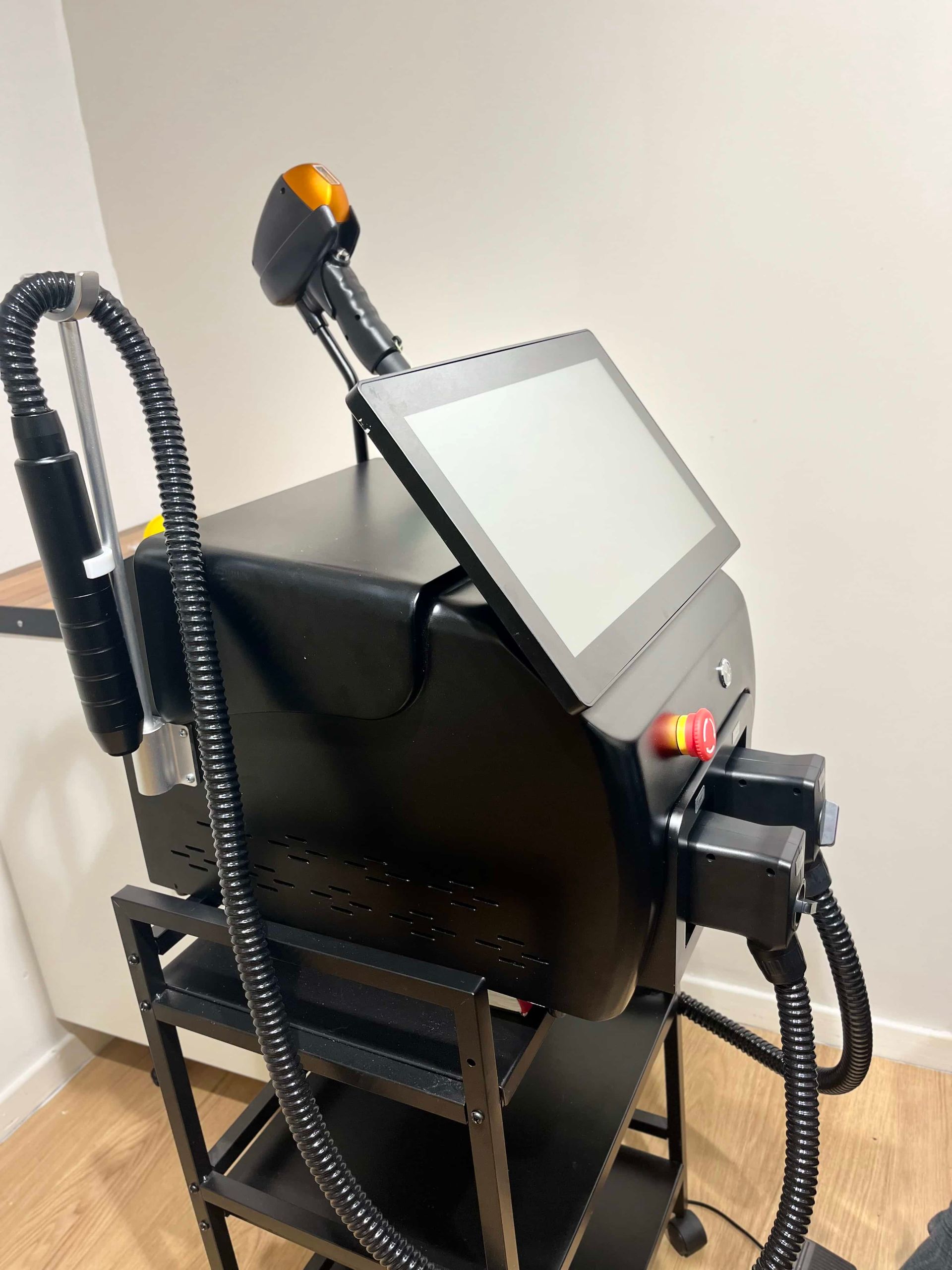 A black laser aesthetic machine with a touchscreen interface and handheld applicator sits on a rolling cart.