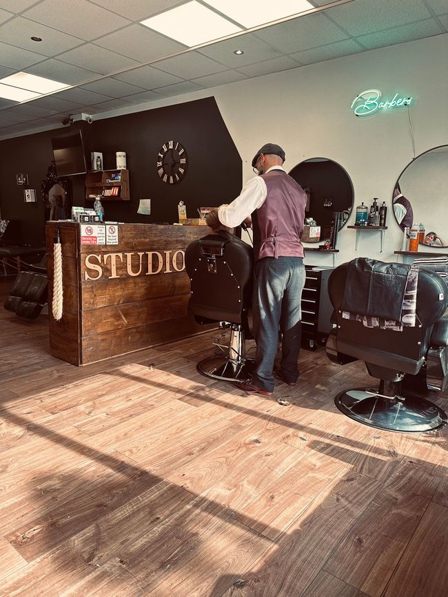 A person wearing a vest and cap stands by a barber chair in a studio, facing a wooden reception desk.