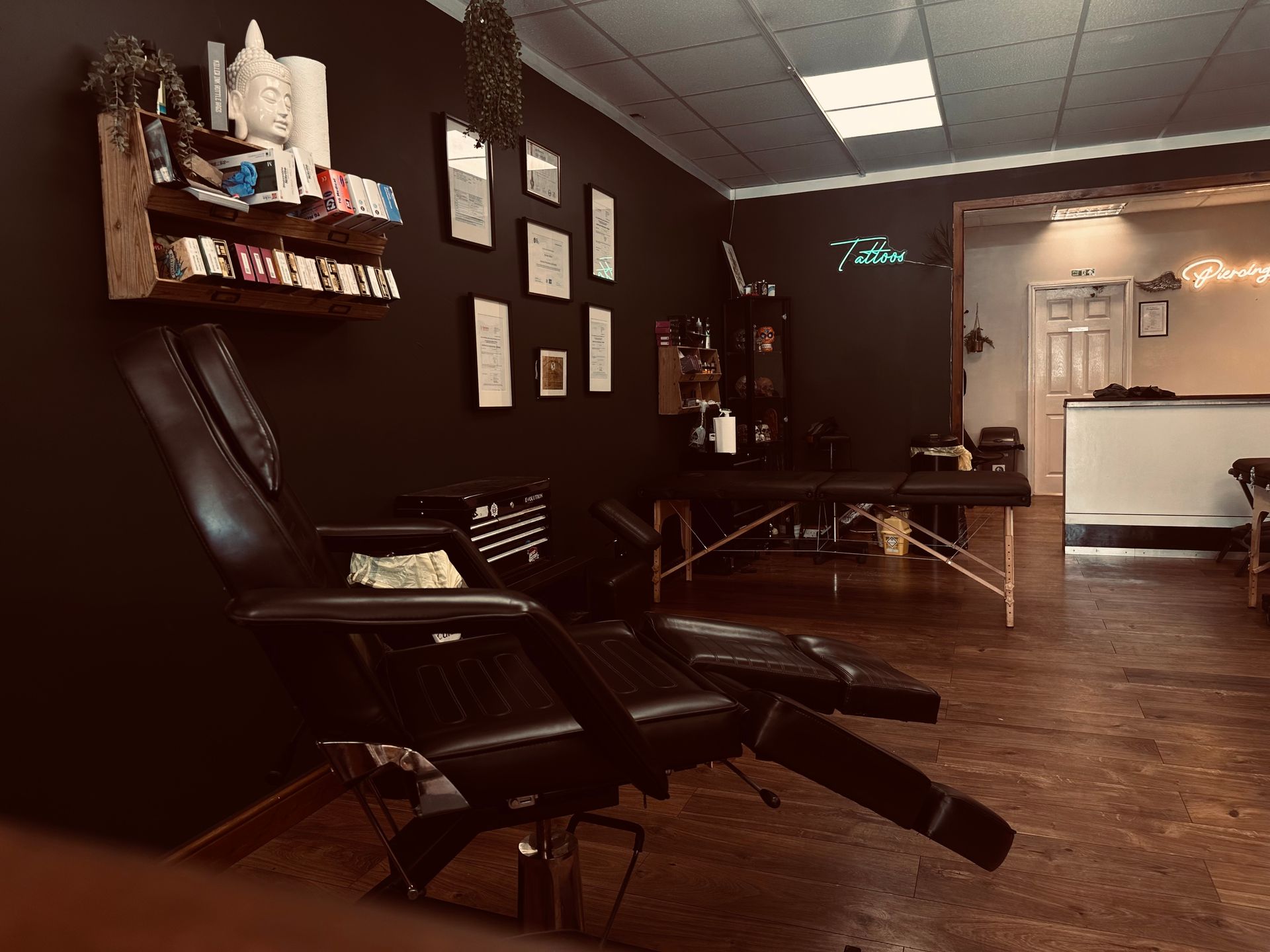 A black-walled tattoo parlor with a reclining chair in the foreground, professional equipment, and a service desk beyond.