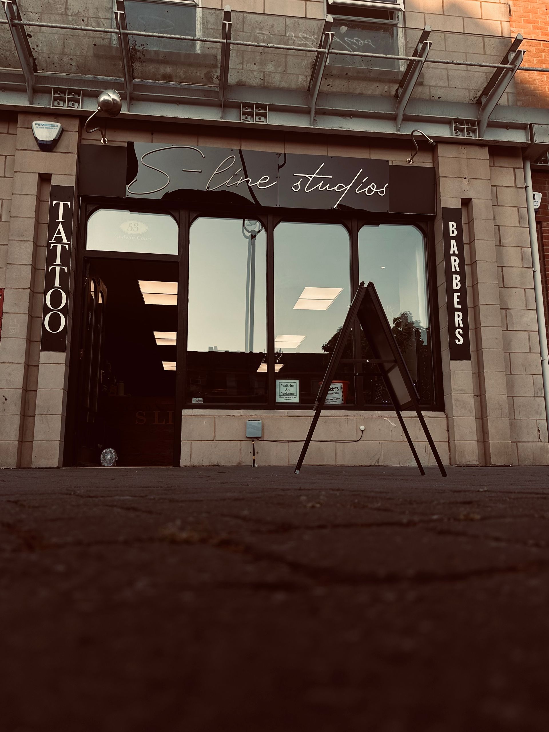 A tattoo and barbershop storefront with a dark sign above the entrance and an A-frame sign on the sidewalk.