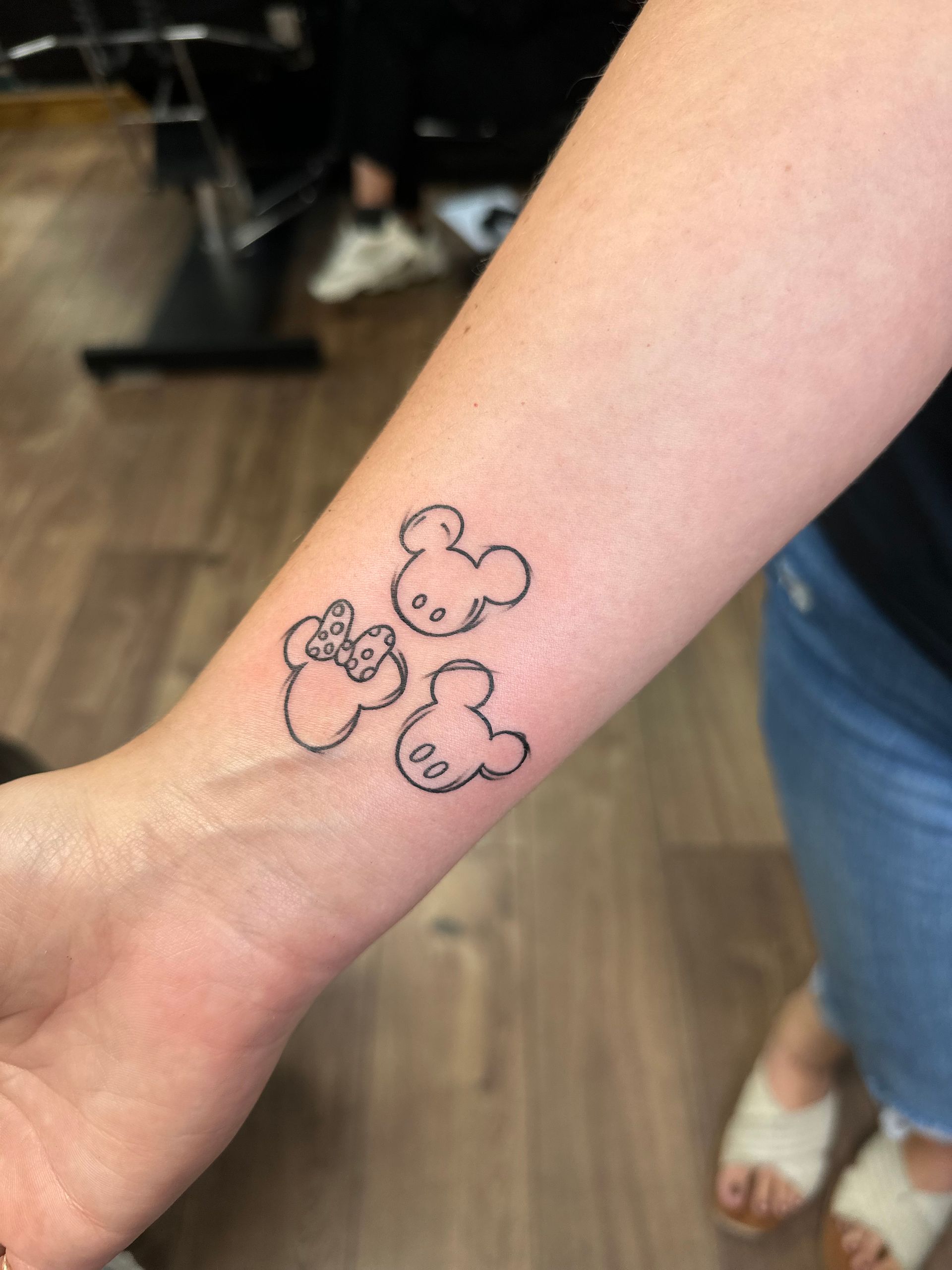 A close-up of a forearm tattoo featuring three black outline Mickey and Minnie Mouse icons in a cluster.